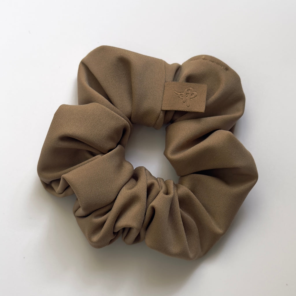 A taupe brown fabric scrunchie with a small tan leather patch featuring an embossed bee logo, shown on a plain white background.
