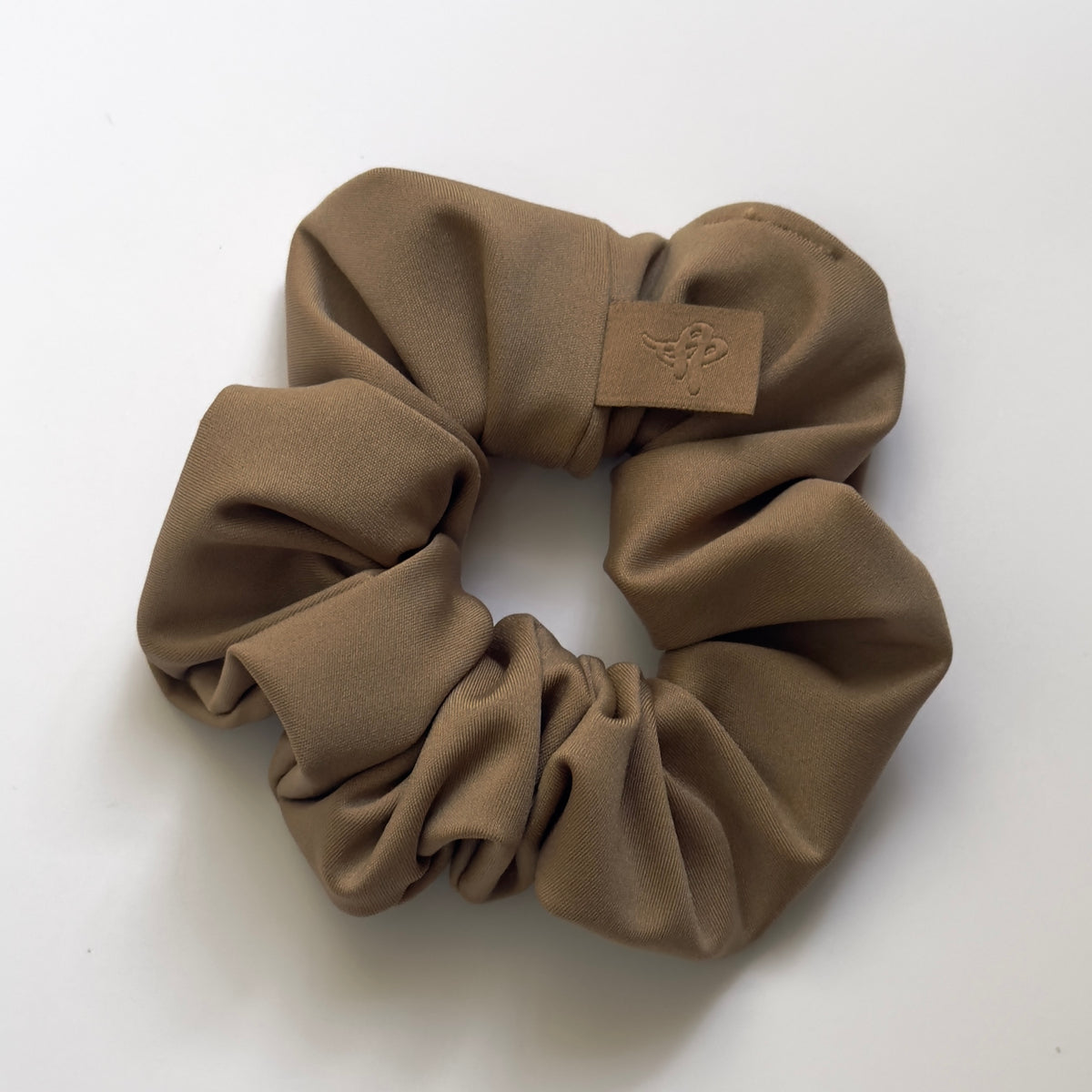 A taupe brown fabric scrunchie with a small tan leather patch featuring an embossed bee logo, shown on a plain white background.