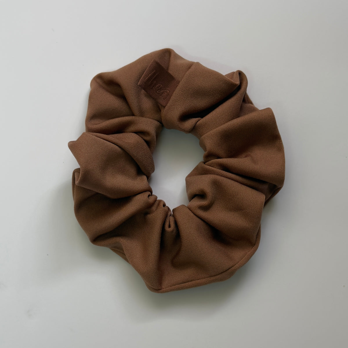 A single brown fabric scrunchie with a gathered elastic band, photographed from above on a plain light background. A small embossed brand tag is visible near the top.