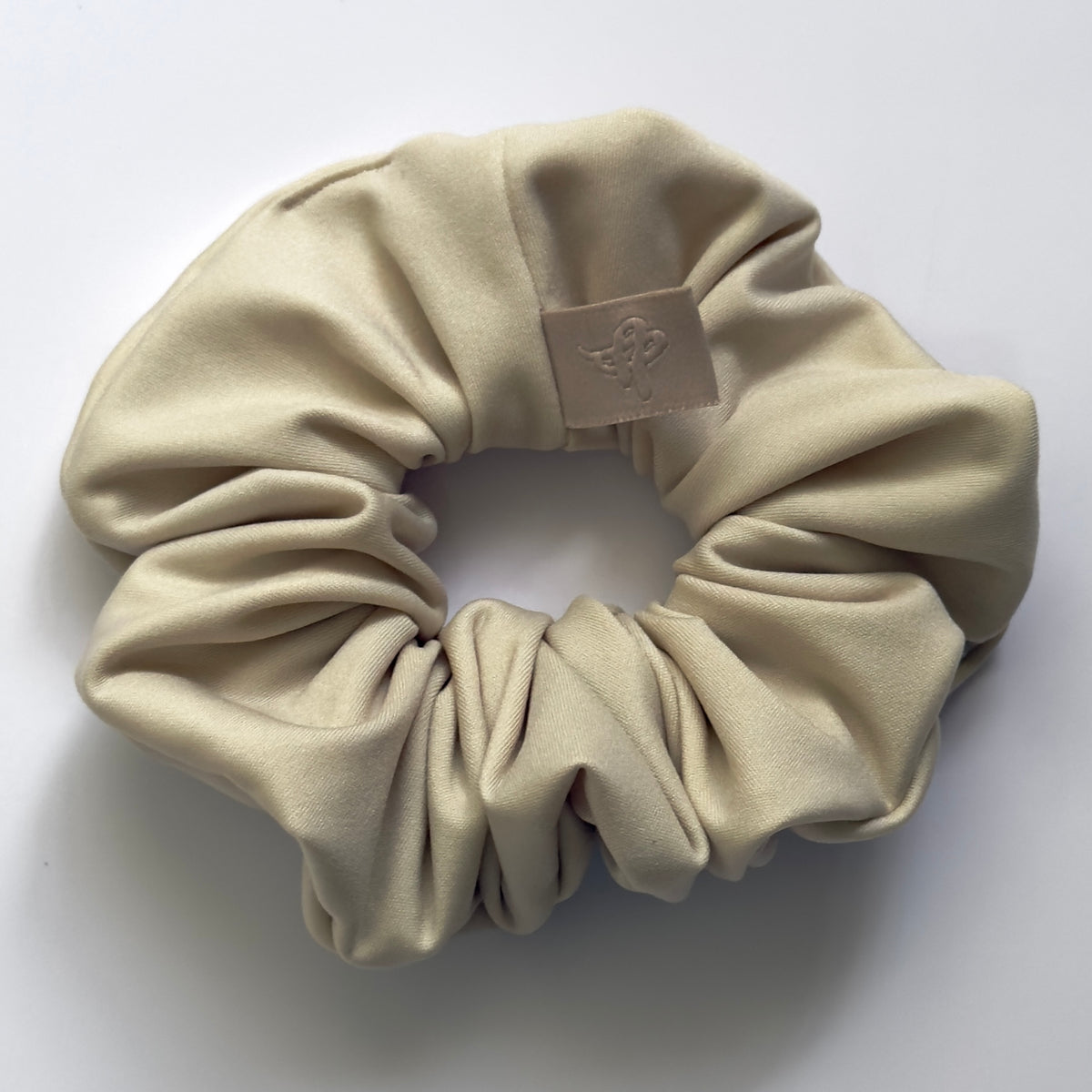 A beige satin hair scrunchie with a small beige leather brand patch on the front, photographed on a clean white background.