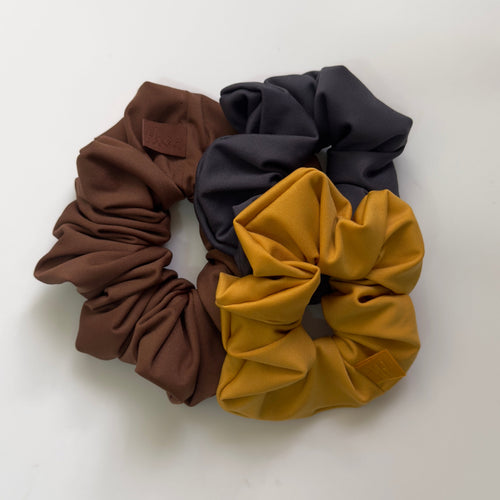 Three fabric hair scrunchies arranged on a plain white background: one medium brown, one dark navy, and one mustard yellow.