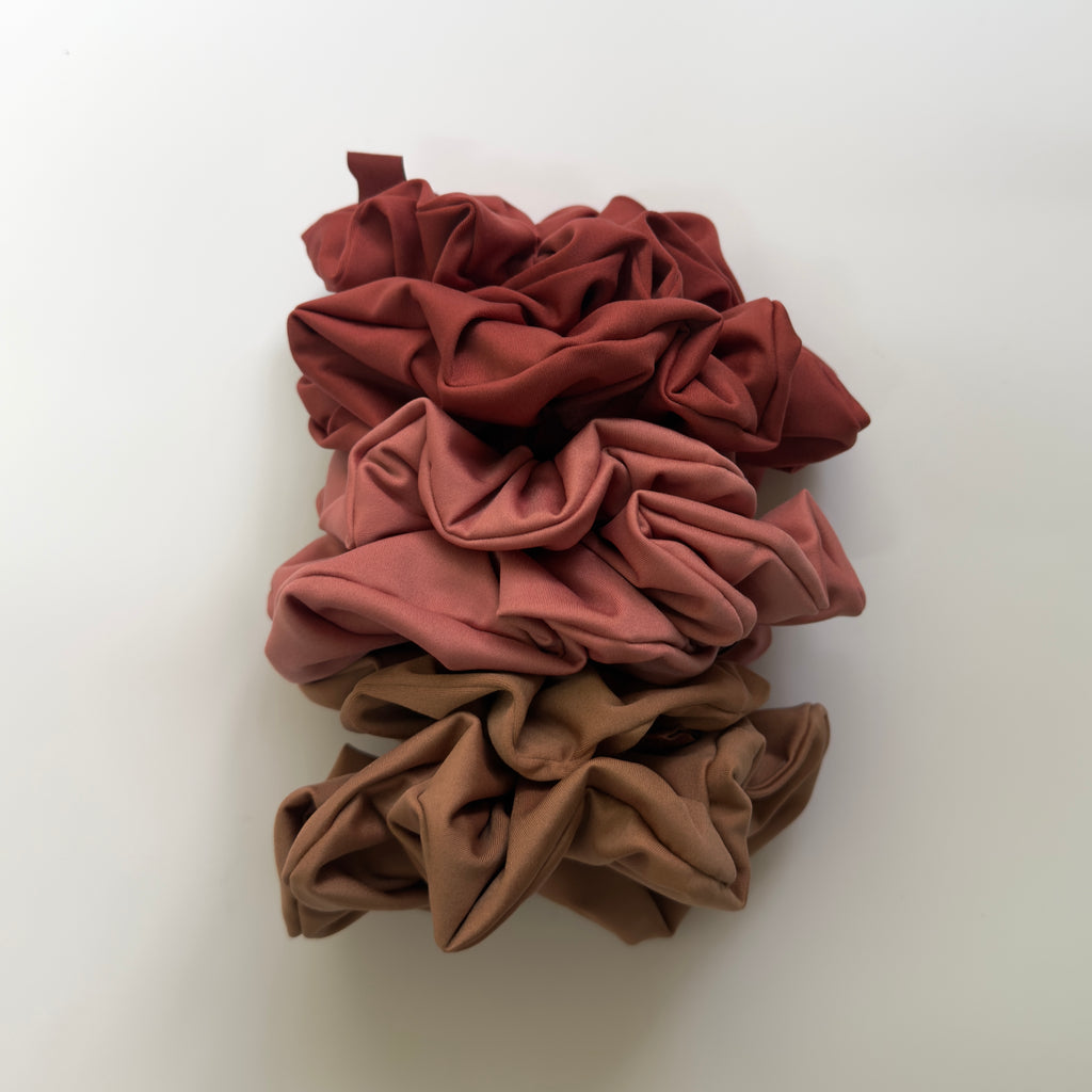 Three fabric hair scrunchies stacked vertically on a plain white background, showing a color gradient from dark terracotta red at the top, through dusty rose pink in the middle, to warm taupe brown at the bottom.