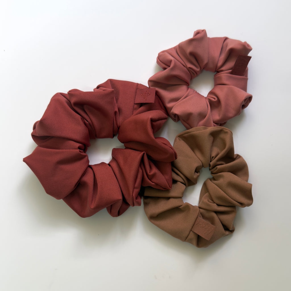 Three fabric hair scrunchies in rust red, dusty rose, and taupe brown arranged on a plain white background.