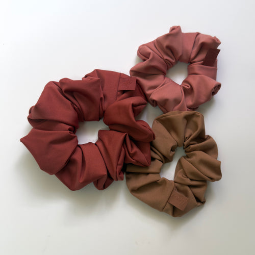 Three fabric hair scrunchies in rust red, dusty rose, and taupe brown arranged on a plain white background.