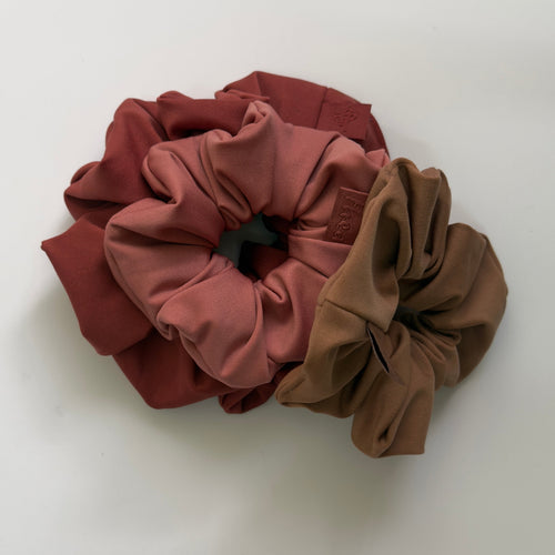Three fabric hair scrunchies in dusty rose, terracotta, and taupe brown, arranged overlapping on a clean white background.