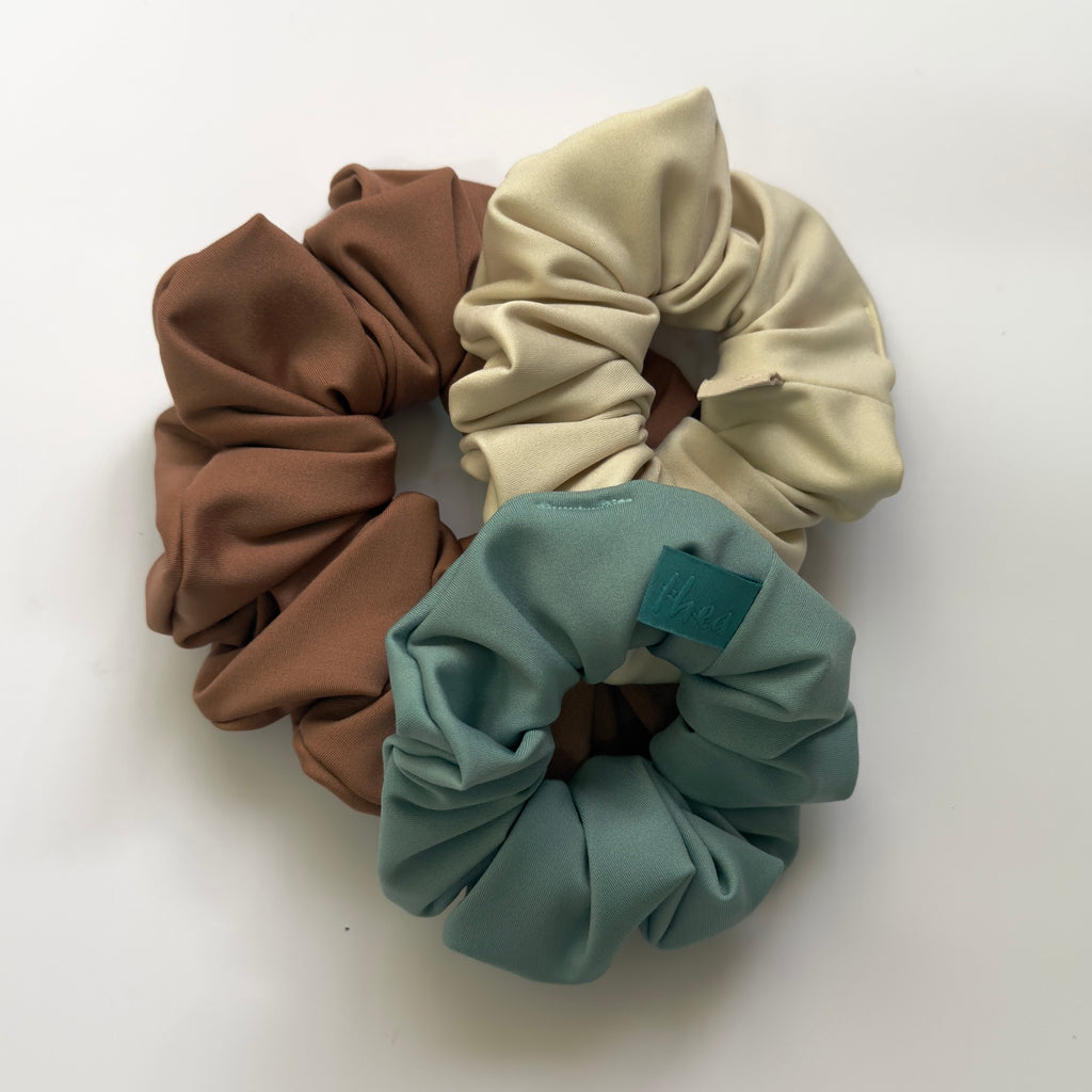 Three fabric hair scrunchies in brown, cream, and teal, arranged on a plain white background.