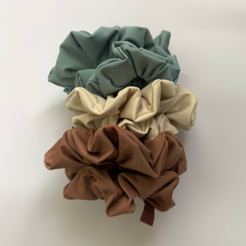 Three fabric hair scrunchies stacked vertically on a plain white background. The top scrunchie is muted sage green, the middle is warm cream, and the bottom is medium brown.