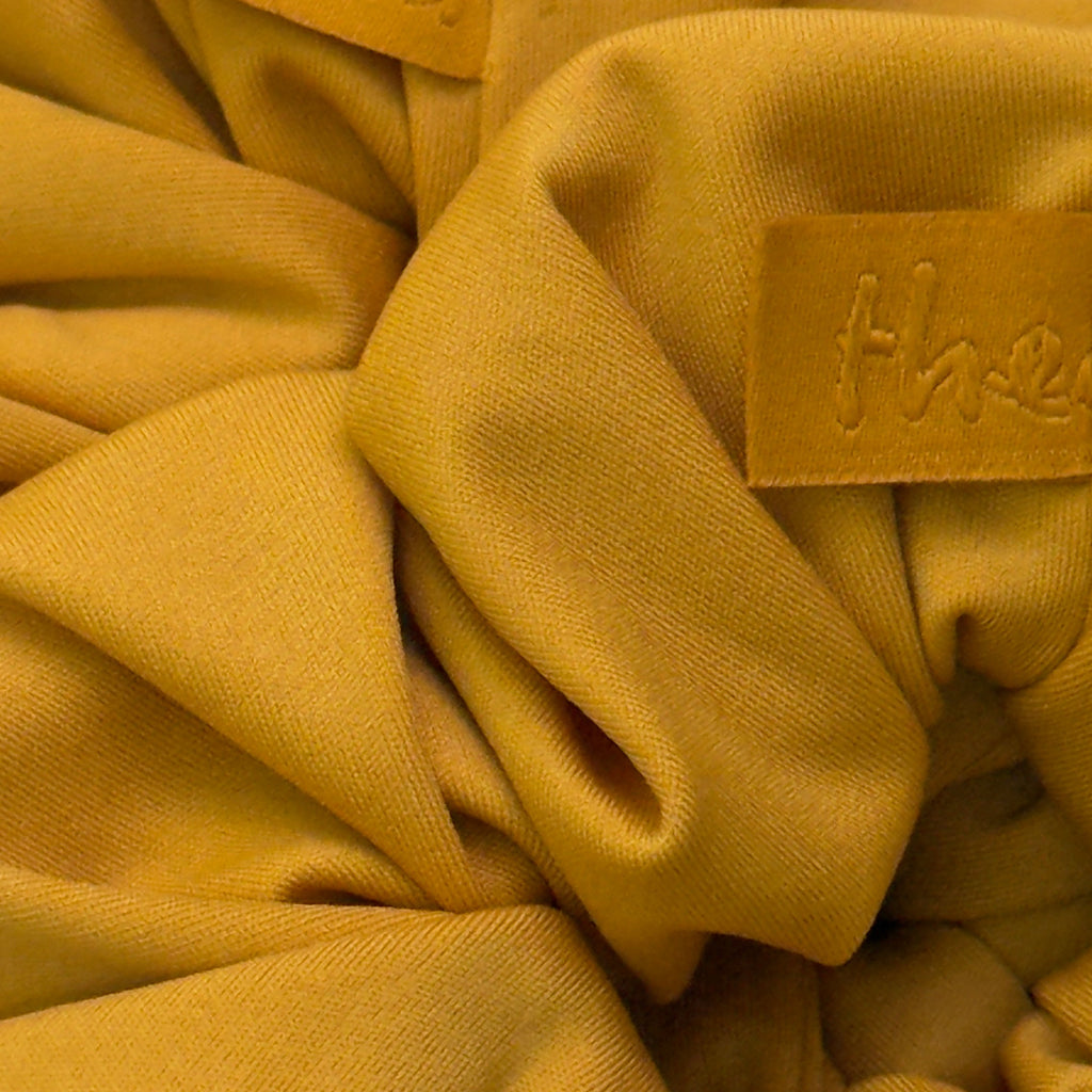 A close-up of gathered mustard yellow fabric with two small tan leather patches embossed with the brand name 'thea'.